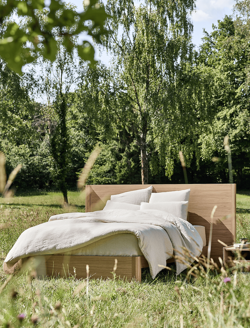 Cama de madera maciza Kipli situada en plena naturaleza, con ropa de cama de lino natural, que ilustra un dormitorio ecológico y un sueño saludable en medio de un entorno verde.