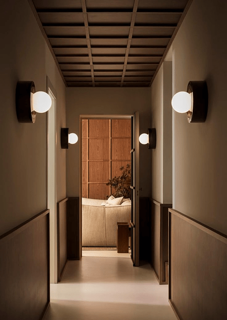 Corredor minimalista iluminado por apliques de parede modernos, que conduz a uma acolhedora sala de estar com acabamentos em madeira e um sofá bege, exemplificando uma decoração de interiores elegante e tranquilizante.