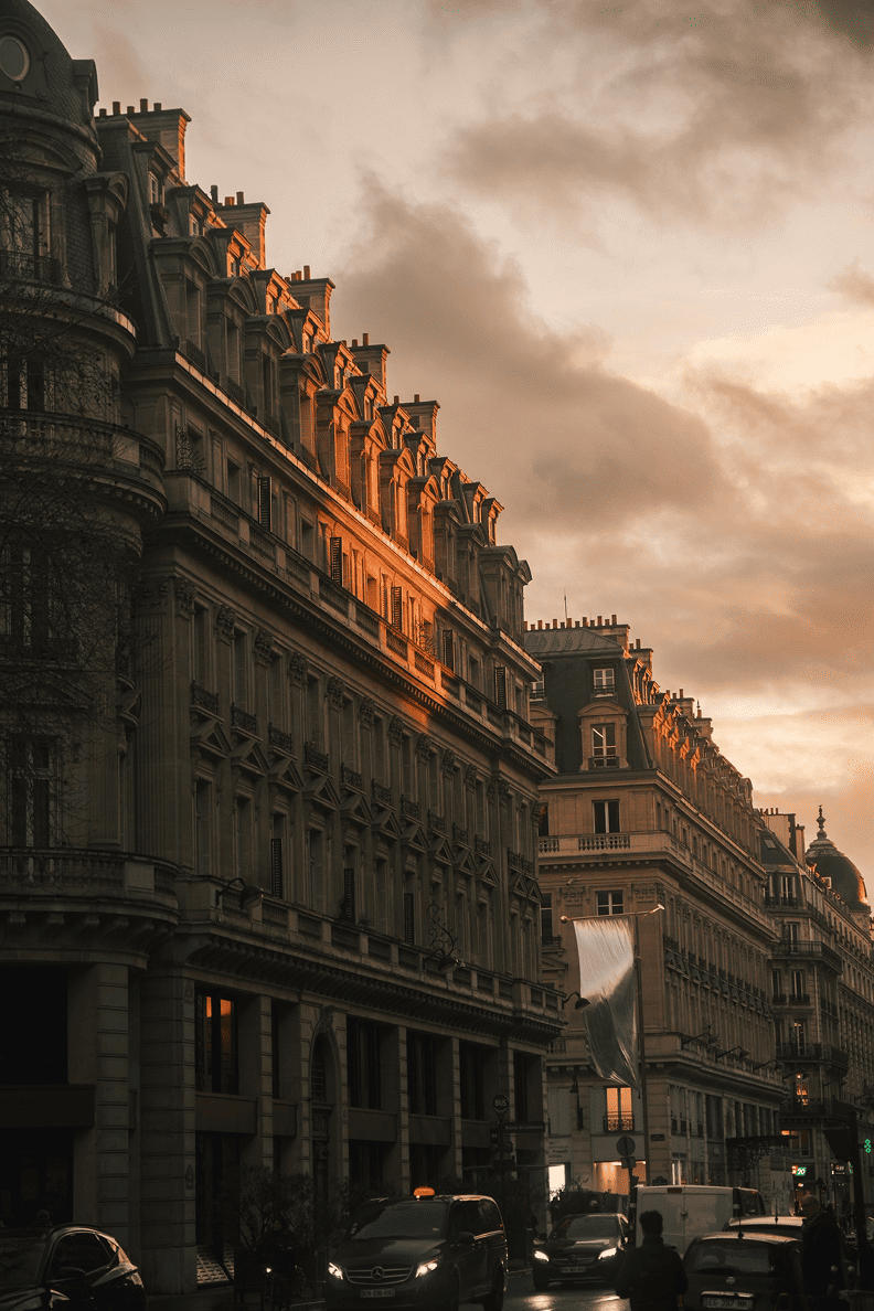 Fachadas haussmannianas em Paris ao pôr-do-sol, com uma luz dourada sobre os telhados e o trânsito à base dos edifícios.
