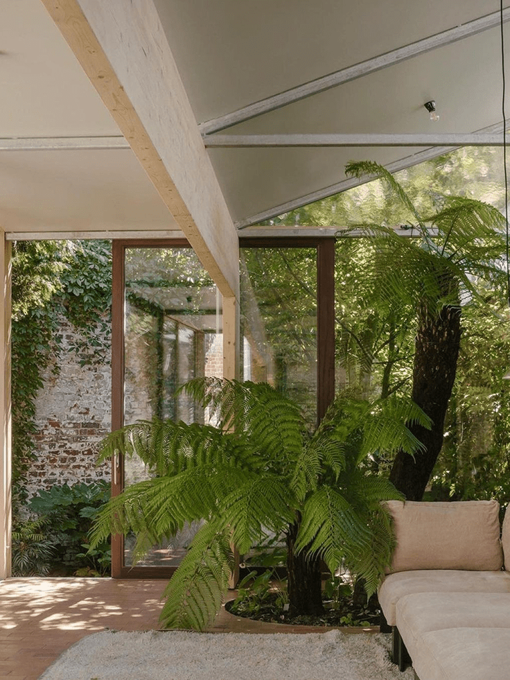 Luminoso salotto dall'arredamento naturale, con grandi piante verdi e una vetrata che si affaccia su un giardino, a creare un ambiente rilassante ed eco-responsabile.